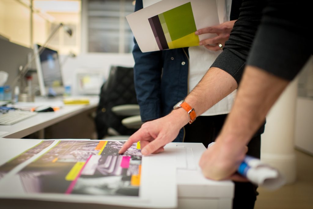 Designers reviewing printed proofs with colorful highlights in a collaborative workspace, emphasizing graphic design for outdoor print campaigns.