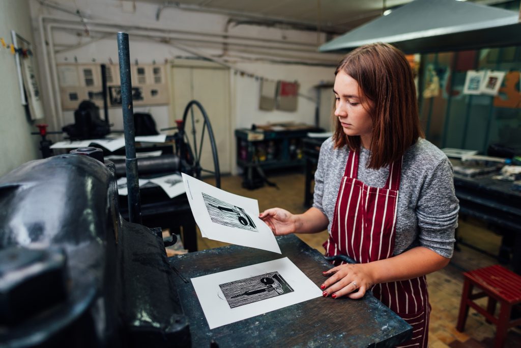 Young woman inspecting printed linocut artwork in a printmaking studio, showcasing the manual printing process related to outdoor advertising campaigns.