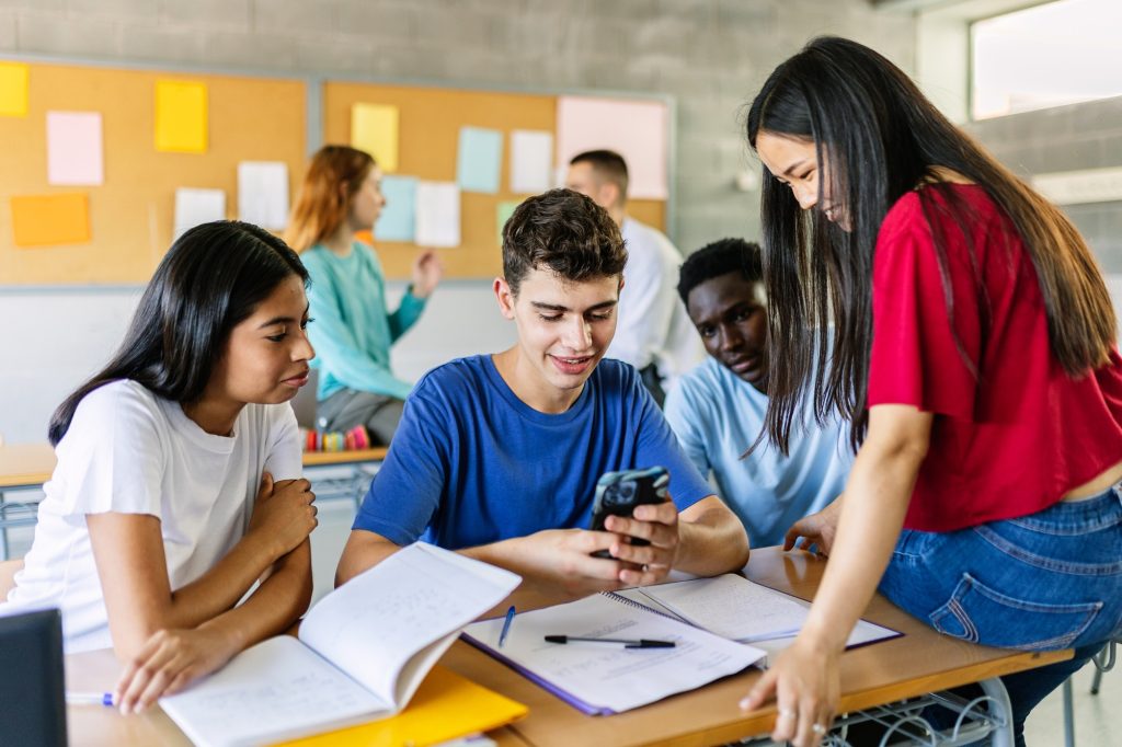 Multiracial group of students engaging with smartphone content while collaborating on schoolwork, highlighting social media's role in fostering connections and community.