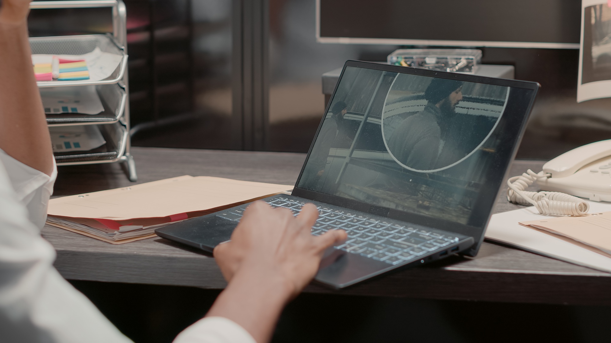 Person using a laptop to study crime case files, surrounded by office materials and a telephone, reflecting the importance of digital tools in modern investigative work.