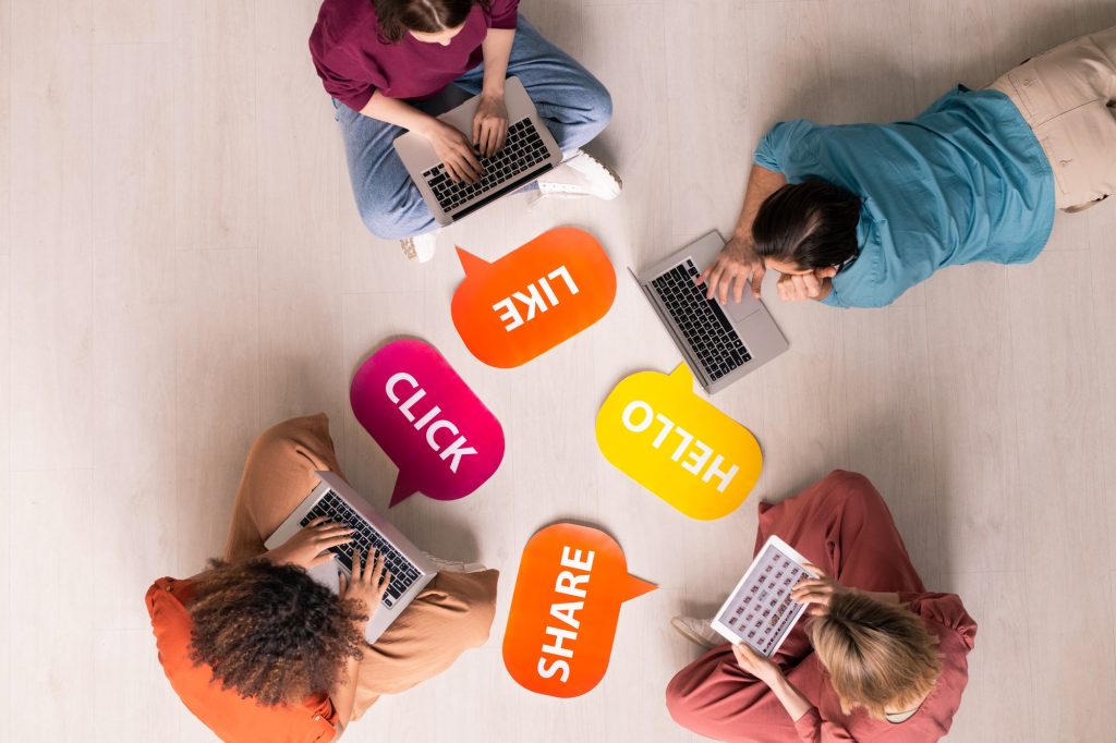 Group of people using laptops surrounded by colorful social media interaction icons labeled "LIKE," "CLICK," "SHARE," and "HELLO," illustrating engagement in digital communication and social media management.