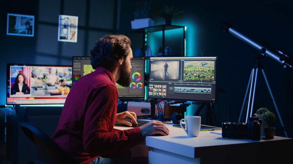 Video editor working on multiple screens with editing software, showcasing video footage, and a coffee mug on desk in a modern production studio.