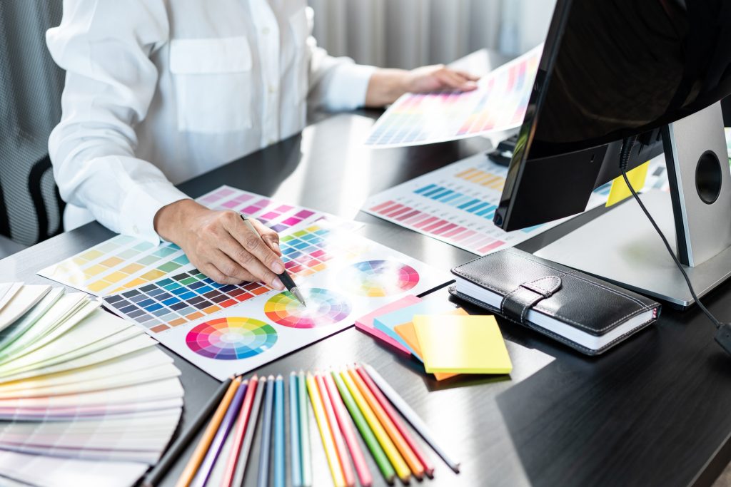 Young graphic designer selecting colors and drawing on a graphics tablet, surrounded by color swatches, sticky notes, and design materials, illustrating the creative process in graphic design.