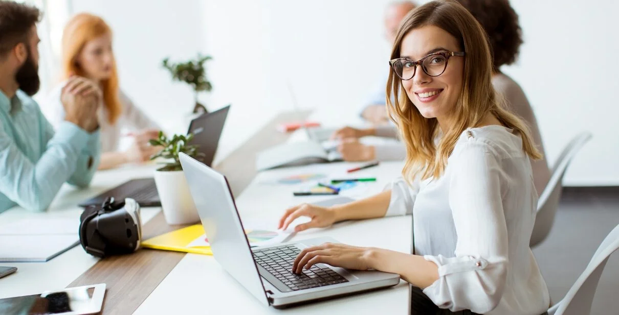 Woman smiling at laptop in a collaborative office setting with colleagues, highlighting digital marketing strategies for business growth.