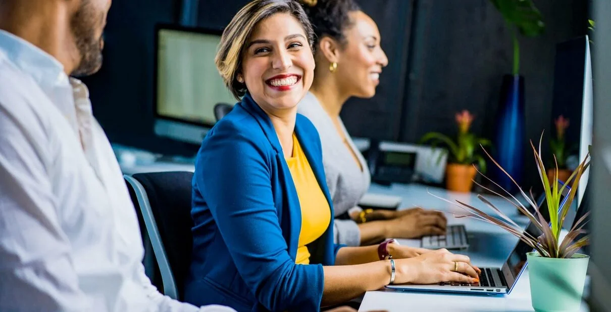 Smiling woman in blue blazer working on laptop in modern office with colleagues and plants, illustrating MediaTamer's digital marketing environment.