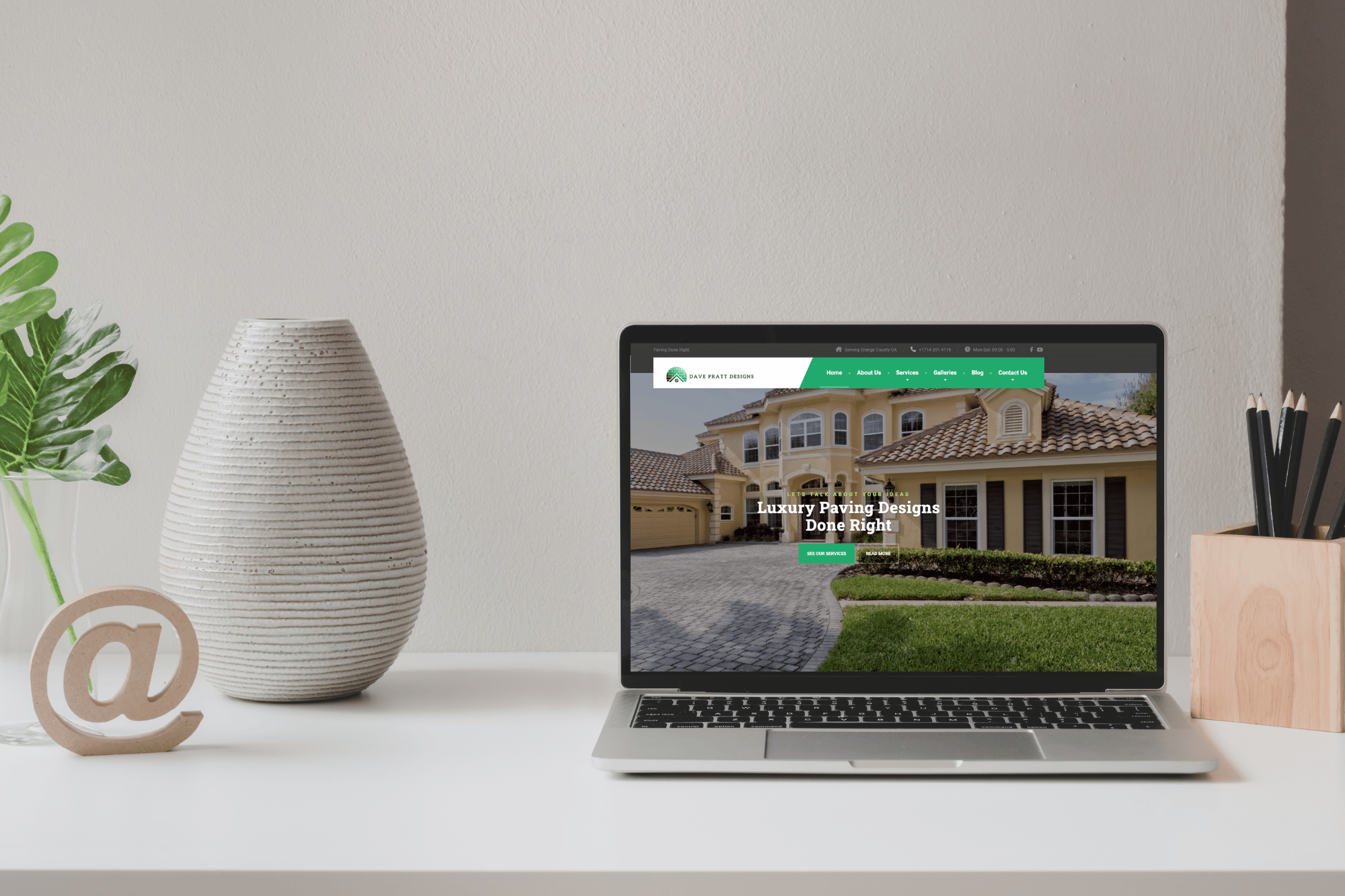 Laptop displaying a luxury paving design website, featuring a modern home exterior, with decorative plant and pencil holder on a clean desk.